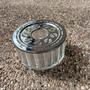 May include: A silver and clear glass tea candle holder with a decorative design. The holder has a round base and a circular top with a cutout design.