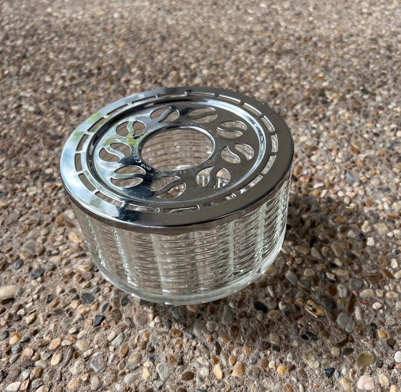 May include: A silver and clear glass tea candle holder with a decorative design. The holder has a round base and a circular top with a cutout design.