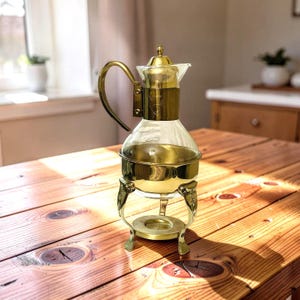 May include: A vintage brass and glass coffee server. It features a clear glass carafe and a brass warming stand. The server has a curved handle and a decorative lid. The stand has ornate feet and a circular base.