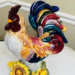 May include: A ceramic rooster figurine with a colorful design. The rooster has a red comb and wattle, a yellow beak, and a white body. The tail feathers are blue, green, red, and yellow. The base features sunflowers and a ladybug.