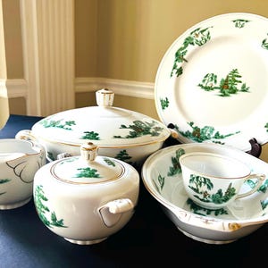 Op de afbeelding: Een set witte porseleinen schalen met groene en gouden accenten. De set bevat een grote serveerschaal met deksel, een kleinere schaal, een roomkan, een suikerpot en een dinerbord. De schalen zijn versierd met een delicaat bloemmotief met groene bladeren en bloemen.