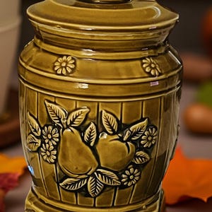 May include: A vintage olive green ceramic jar with a lid, featuring a decorative relief of pears, apples, and flowers. The jar has a barrel-like design with horizontal ridges and a square base. A small teapot-shaped handle sits atop the lid.
