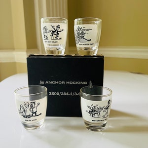 May include: Set of four clear glass shot glasses with black and white cartoon illustrations. The illustrations depict humorous scenes with the text "Who's got your eye?", "Drinks are on the house", "Down the hatch!" and "Here's looking at you, kid". The glasses are packaged in a black box with the text "By Anchor Hocking 3500/384-1/3-9".
