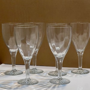 May include: Six clear glass wine glasses with a fluted design on the base. The glasses are empty and arranged in a row on a white surface.