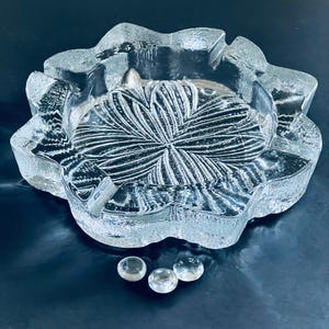 Puede incluir: Un cenicero de vidrio transparente con un diseño de estrella. El cenicero tiene un borde ondulado y está sobre una superficie azul oscuro.