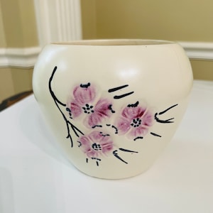 Puede incluir: Un macetero de cerámica blanco con un diseño floral. El macetero presenta una rama con flores rosas y contornos negros.