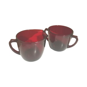 Könnte beinhalten: Zwei rote Glas-Sahnegießer mit Henkeln. Die Gießer sind aus rubinrotem Glas und haben eine abgerundete Form.
