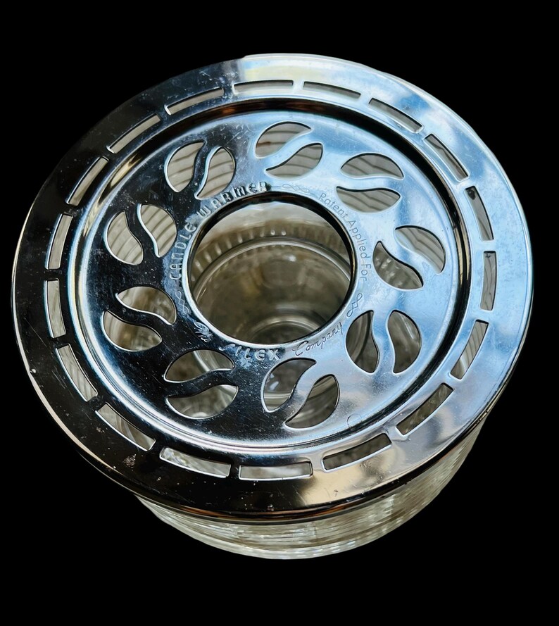 May include: Silver candle warmer with a glass base and a decorative metal top with a floral design. The top has the text "Candle Warmer" and "Patent Applied For" on it.