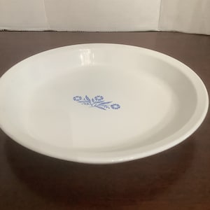 May include: White pie plate with a blue floral design. The design features a cluster of blue flowers and leaves.