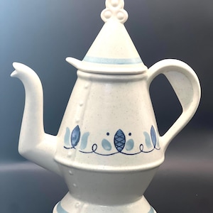 May include: Cream-coloured ceramic teapot with a conical lid and a curved spout. The teapot features blue floral designs and a blue stripe around the base and lid. The handle is a curved loop. The lid has a decorative knob.
