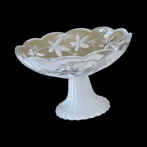 Puede incluir: Un cuenco de pedestal de vidrio transparente con borde festoneado y diseño floral grabado. El cuenco se asienta sobre una base de pedestal blanca y acanalada. Adecuado para servir postres o exhibir objetos decorativos.