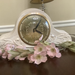 May include: White mantle clock with gold accents and a floral design. The clock face has black hands and Roman numeral markings. The clock is sitting on a dark brown surface with pink flowers.