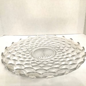 May include: A clear glass serving plate with a scalloped edge and a diamond pattern. The plate has a smaller, circular center piece with a starburst design.
