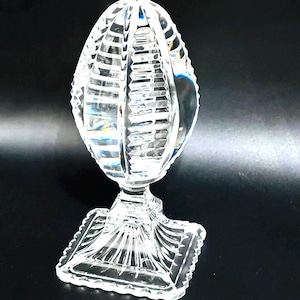 May include: A clear, cut-glass decorative egg-shaped object on a square base. The egg has vertical and horizontal cut lines, creating a textured surface. The base has a detailed, geometric design. The object is displayed against a black background.
