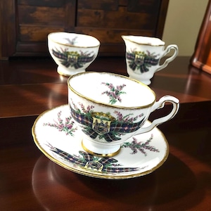 May include: A white teacup and saucer set with gold trim, featuring a plaid ribbon design with a crest and floral accents. The set includes a creamer and two small bowls. The teacup is on a saucer, and the set is displayed on a dark wooden surface.