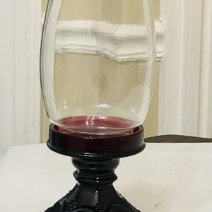 Puede incluir: Un portavelas de cristal transparente con la parte superior acampanada, sobre una base de pedestal roja oscura y negra. La base tiene un diseño decorativo tallado. El portavelas está diseñado para uso decorativo.