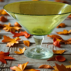 Può includere: Una ciotola verde in vetro con piede e un bacino ampio e poco profondo. La ciotola poggia su uno stelo e una base in vetro trasparente. Lo sfondo presenta foglie autunnali nelle tonalità dell'arancione e del rosso, sparse su una superficie di legno.