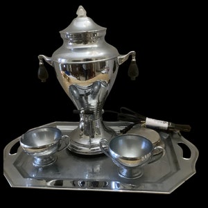 Puede incluir: Una cafetera eléctrica de plata con dos tazas a juego en una bandeja de plata. La cafetera tiene un cable negro y una perilla negra en el lateral.