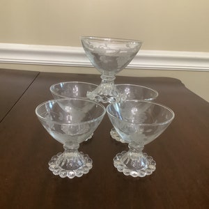May include: A set of five clear glass dessert bowls. Each bowl has a stemmed base with a ring of small, round details. The bowls are etched with a floral design. The bowls are arranged on a dark brown surface.
