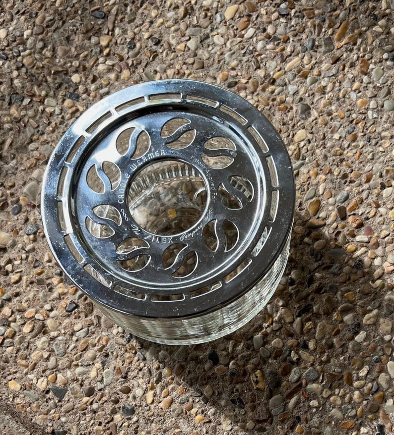May include: Silver candle warmer with a glass base and a decorative design of leaves. The warmer has the text "Candle Warmer" printed on the side.