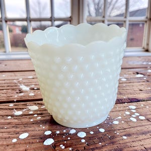 May include: A cream-coloured milk glass vase featuring a raised hobnail texture across its exterior surface. The vessel has a wide, circular opening with a scalloped rim edge and a tapered base, showcasing a translucent quality in the glass.