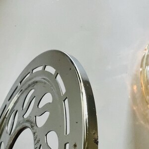 May include: A silver-colored metal bicycle chainring with a decorative design. The chainring has a circular shape with a central hole and a series of cutouts around the edge.