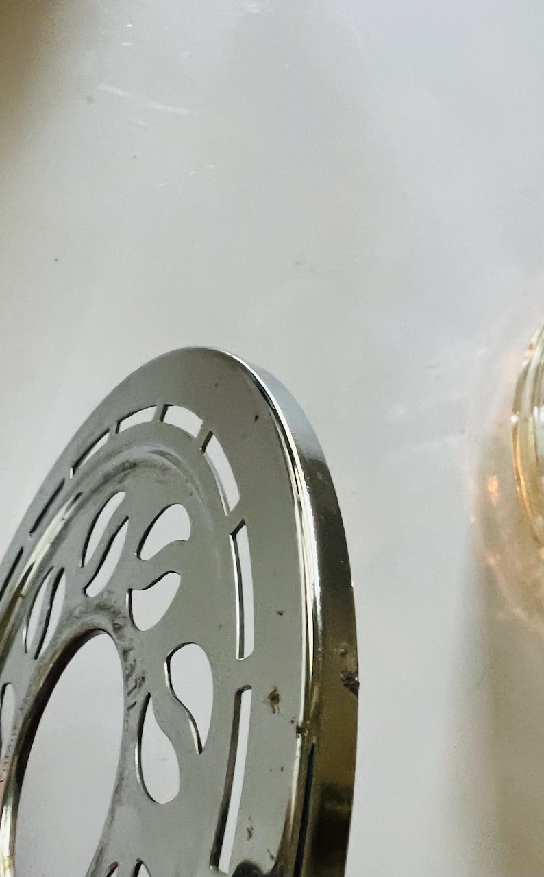 May include: A silver-colored metal bicycle chainring with a decorative design. The chainring has a circular shape with a central hole and a series of cutouts around the edge.