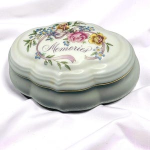 Peut inclure: Une boîte à bijoux ovale en porcelaine avec un bord festonné et une base blanche. Le couvercle présente un motif floral avec des roses roses et jaunes, des myosotis bleus et un ruban violet. Le mot Memories est écrit en cursive sur le couvercle.