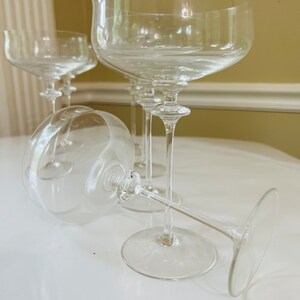 May include: Four clear glass champagne coupes with a wide bowl and a long stem. The glasses are arranged in a group on a white surface.