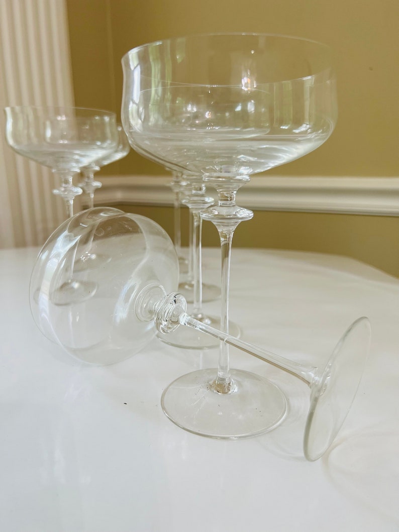 May include: Four clear glass champagne coupes with a wide bowl and a long stem. The glasses are arranged in a group on a white surface.