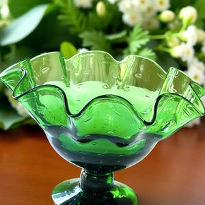 Peut inclure: Un bol en verre vert avec un bord ondulé et un pied. Le bol présente une texture à bulles et un contour vert foncé sur le bord. En arrière-plan, des fleurs blanches et du feuillage vert.