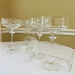 May include: Six clear glass champagne coupes with long stems. The glasses are arranged in a row on a white surface.