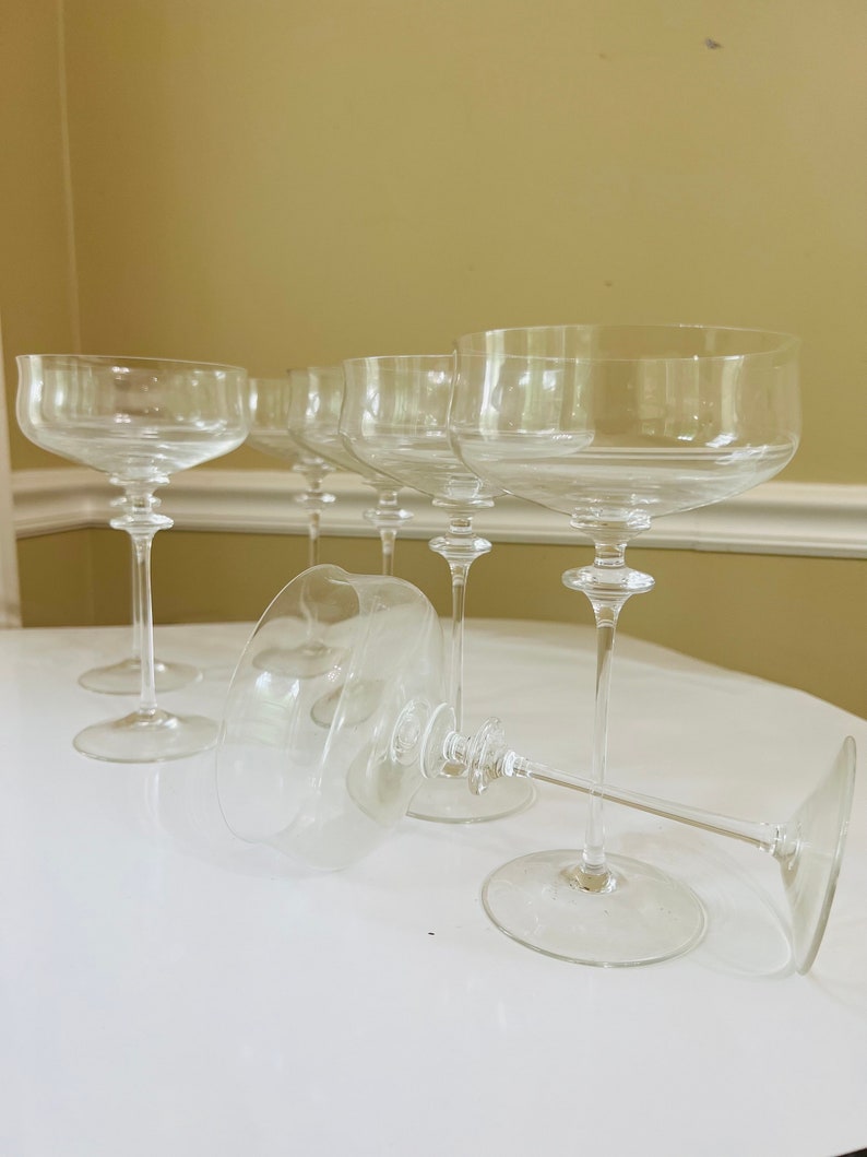 May include: Six clear glass champagne coupes with long stems. The glasses are arranged in a row on a white surface.