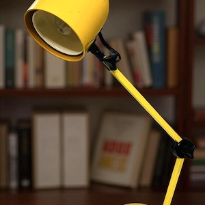 May include: A bright yellow desk lamp with a rounded shade and base. The lamp has a long, adjustable arm and a black joint. The lamp is on a wooden surface, with a bookshelf in the background.