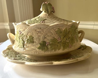 May include: A ceramic serving dish with a lid, featuring a detailed design of woven wicker and green leaves with pink berries. The dish is in a cream color with green and pink accents.