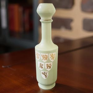 May include: A pale green decorative bottle with a bulbous top and a slender neck. The bottle features gold and white heraldic crests. The bottle is displayed on a dark wooden surface, with a blurred background.