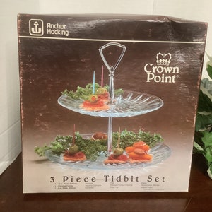 Puede incluir: Un juego de servir de tres piezas de vidrio con un soporte cromado. El juego incluye dos platos de vidrio transparente con un patrón decorativo y un soporte cromado con un asa. El juego está etiquetado como "Crown Point" y "3 Piece Tidbit Set".