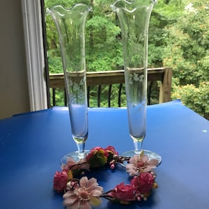 May include: Two clear glass bud vases with a ruffled top and etched floral designs. The vases are sitting on a blue surface with a pink flower crown.