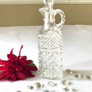 May include: A clear glass bottle with a diamond pattern and a stopper. The bottle has a handle and is sitting on a white surface with clear glass stones.