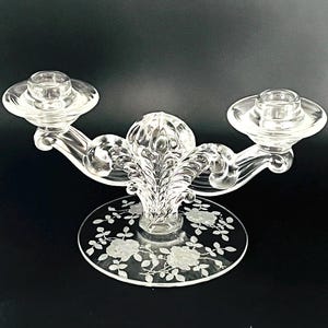 Puede incluir: Candelabro de cristal transparente con dos brazos y un elemento decorativo central. La base presenta diseños florales grabados. Los candelabros son redondos y tienen un pequeño borde para sujetar las velas. El diseño general es elegante y decorativo.