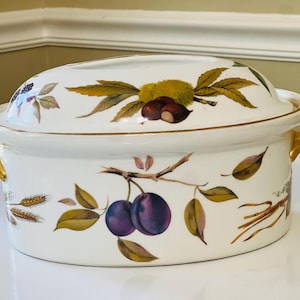 Puede incluir: Plato de cerámica ovalado blanco con borde y asas doradas. El plato está decorado con un patrón floral con ciruelas, hojas y espigas de trigo.