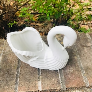 Puede incluir: Macetero de cerámica blanca en forma de cisne con un diseño detallado. El cuello del cisne está curvado y el macetero tiene una abertura ancha.