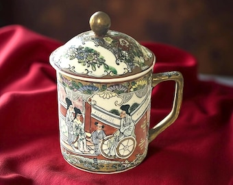 Puede incluir: Una taza de cerámica con tapa, con un diseño de inspiración asiática. La taza tiene un asa dorada y un botón dorado en la tapa. La obra de arte representa figuras con vestimenta tradicional. El fondo es rojo oscuro.