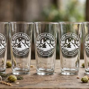 May include: A set of clear glass pint glasses, each featuring a white circular logo that displays the text High Peak Brewing, Est. 2019, Craft Brewery, and a graphic illustration of mountains, pine trees, a star, and hop cones.