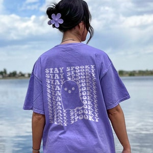 Può includere: Una t-shirt viola a maniche corte con una grafica bianca sul retro. Il design include un'illustrazione centrale di un fantasma circondata dal testo ripetuto STAY SPOOKY disposto in un motivo a griglia ondulato e distorto sul tessuto.