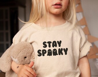 May include: A beige short-sleeved t-shirt featuring the black text STAY SPOOKY with two small ghost illustrations. The shirt is paired with black shorts and is held alongside a tan plush toy. The design is centered on the chest of the garment.