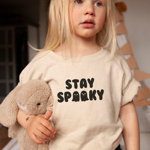 Pu&ograve; includere: Una t-shirt beige a maniche corte con la scritta nera STAY SPOOKY e due piccole illustrazioni di fantasmi. Il capo &egrave; abbinato a pantaloncini neri e tenuto insieme a un peluche beige. Il disegno &egrave; posizionato al centro sul petto della maglia.