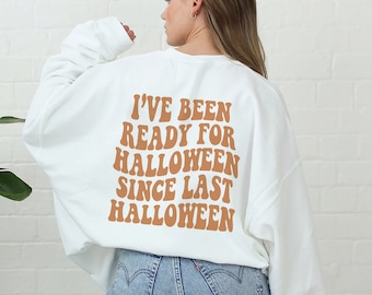 May include: A white crewneck sweatshirt featuring the text I'VE BEEN READY FOR HALLOWEEN SINCE LAST HALLOWEEN printed in a large, wavy, light brown font on the back. The sweatshirt is paired with light blue denim jeans.