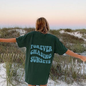 May include: A dark green short-sleeve t-shirt featuring the text "FOREVER CHASING SUNSETS" printed in a light blue, wavy, retro-inspired font on the back. The garment has a crew neckline and a loose, relaxed fit, displaying the graphic clearly.