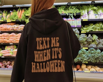May include: A black hooded sweatshirt featuring the brown text "TEXT ME WHEN IT'S HALLOWEEN" printed on the back in a bold, wavy font. The hoodie has long sleeves, a hood, and a relaxed fit, shown from the back view with a black shoulder bag.